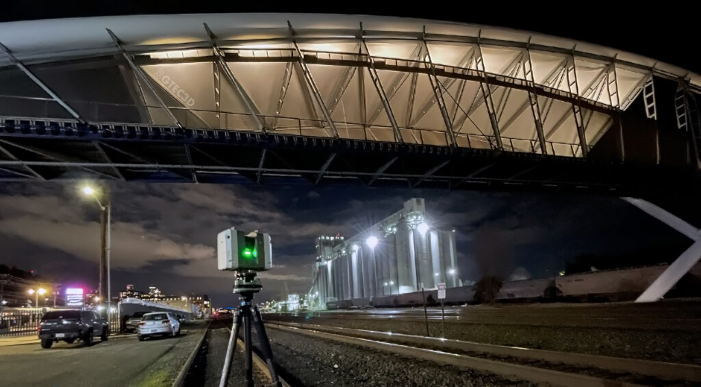 A GTEC3D technician operates a red Leica RTC360 laser scanner on a tripod, capturing 3D data of industrial infrastructure near a waterfront.