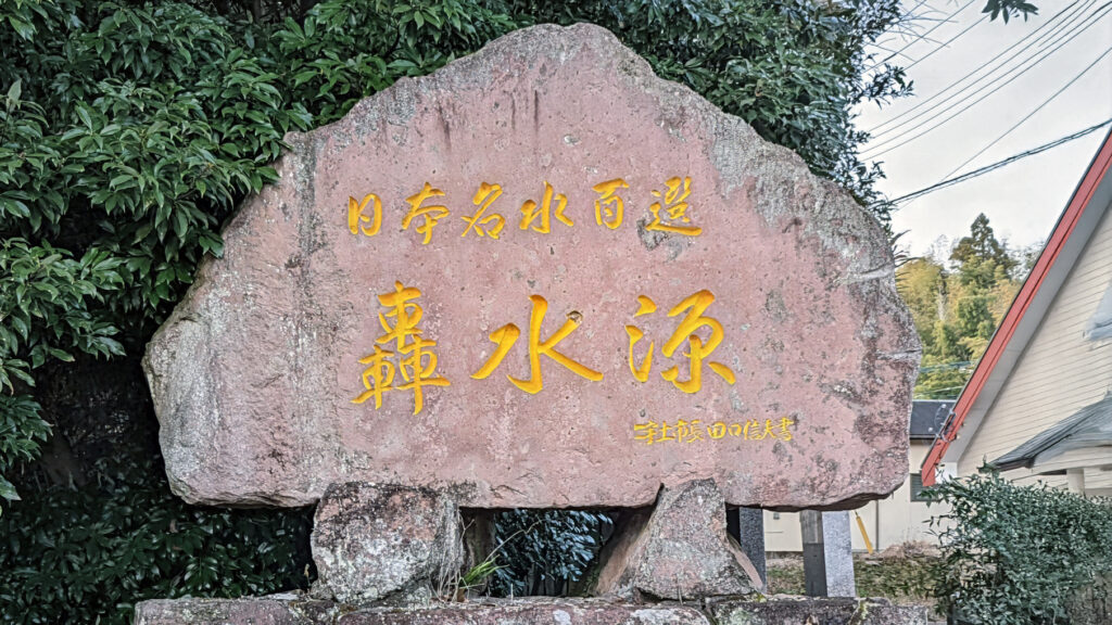 An old, weathered rock sign with Japanese characters for "Todoroki Suigen" (轟水源) in Kumamoto, Japan. The sign stands near a natural spring source, possibly as part of a 3D scanning project by GTEC3D.