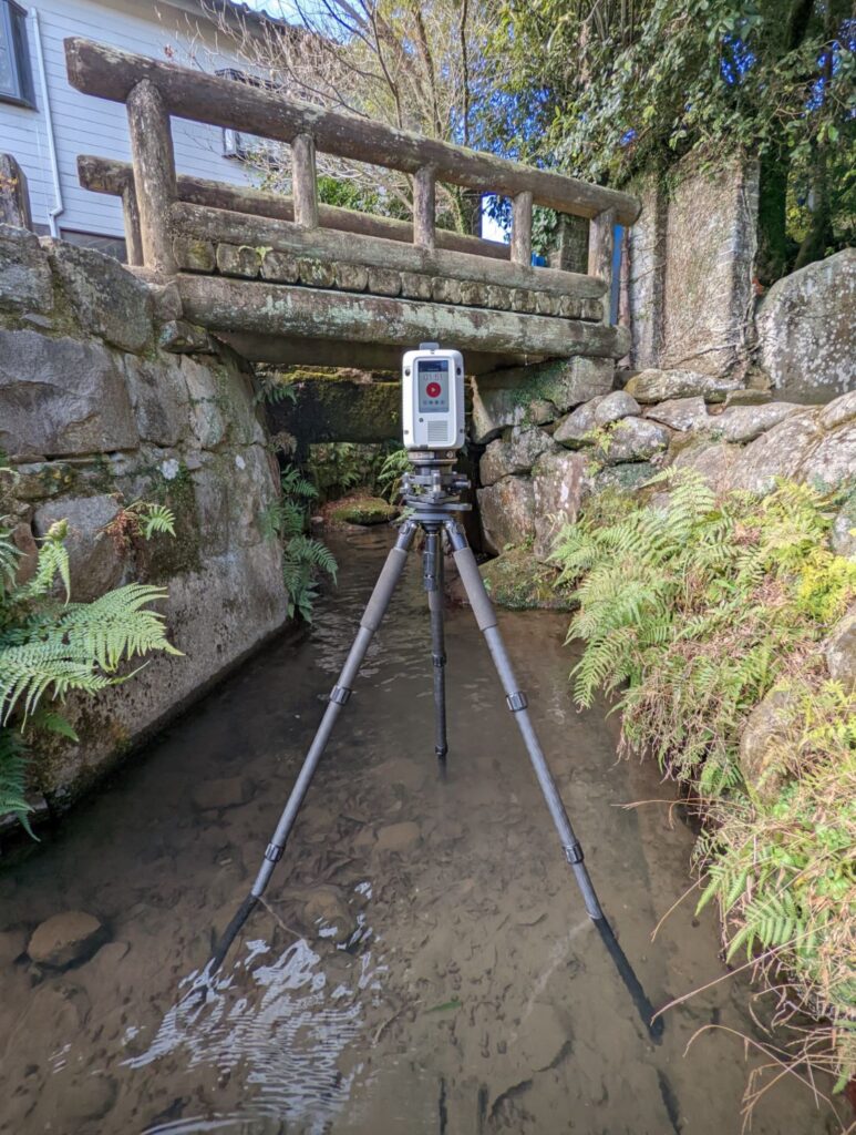 A Leica RTC360 3D laser scanner positioned in the shallow river at Todoroki Suigen captures data of the historic Japanese bridge, with an elderly Japanese man observing nearby.